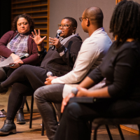 A woman holding a microphone sits in a group of people and speaks passionately.