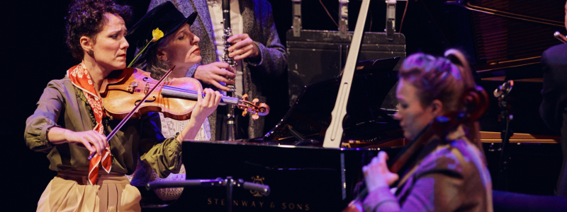 A group of musicians play on a crowded stage. There is a woman playing a violin, a woman playing the cello, and a man playing the clarinet in the background.