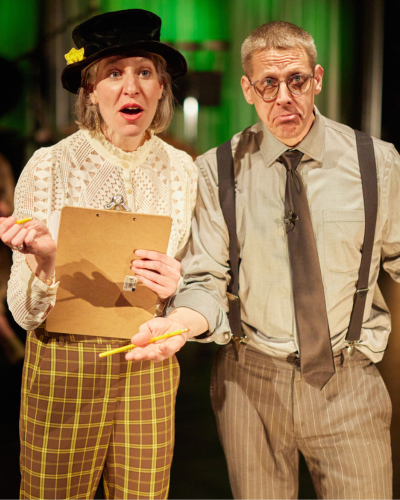 A woman in a white top, a black top hat, and brown and yellow checkered pants holds a clipboard. She is standing to a man wearing suspenders and a brown tie. He is shrugging.