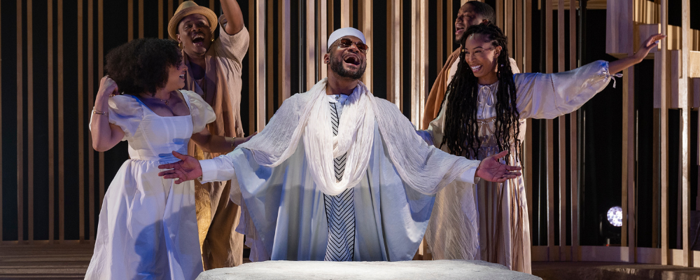 Five individuals stand around a large, flat-topped, light gray rock. To the left, there is a woman in a flowy white dress and a man dressed in beige behind her. In the center is a man in white robes, a white cap, a white scarf, and small black glasses. Next to him is a woman in a beige and white dress, and there is a man in beige behind her. All are singing, with hands raised, looking happy.