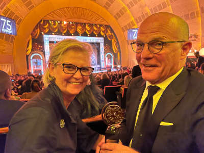 Two people hold a Tony Award and grin. They are overjoyed.