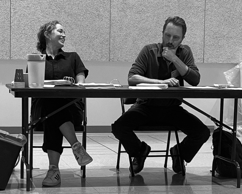 A man and a woman read scripts seated behind a table. The woman is laughing and the man looks perplexed.