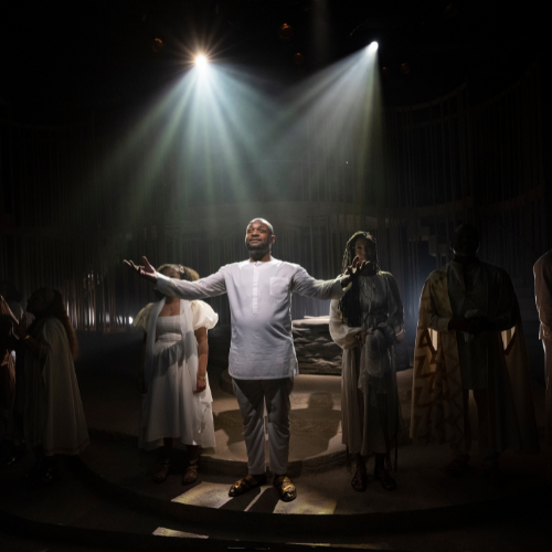 A man in white robes stands on a dark stage as light shines down on him.