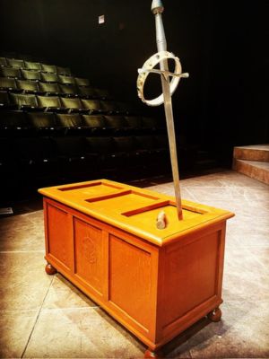 A wooden chest onstage. There is a sword that's been stabbed into it, and a crown resting on the sword.