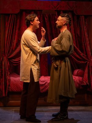 Two men stand in front of a bed with their hands on each others' chests.