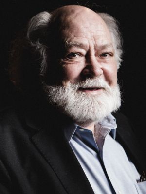 A photograph of the face of Nicholas Rudall, an older white man with white hair and a beard against a dark background. He is looking at the camera and wearing a light blue button down shirt. His mouth is slightly parted, and he is gently smiling.