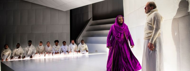 A man in a purple robe turns towards a man in white robes. He looks angry. There is a crowd of people kneeling behind the man in purple.