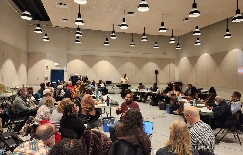 A crowded room of people sitting in chairs. A woman in the background addresses them.