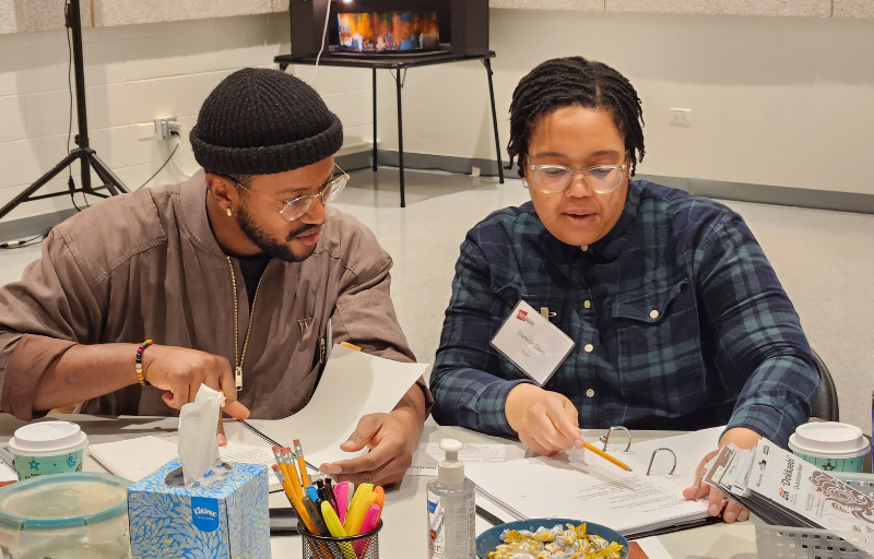 Two people look over a script while sitting at a table.