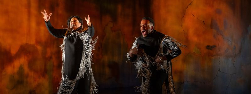 Two people jumping and shouting onstage. They are wearing black robes with silver metallic fringe.
