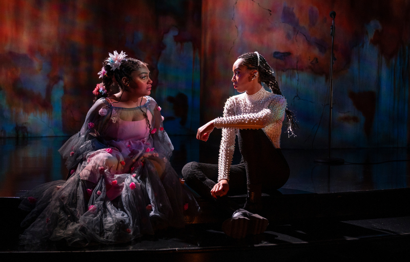 Two women sit on a stage looking at each other.