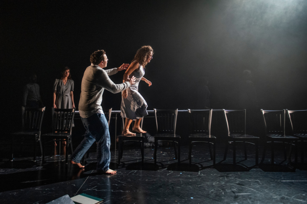 A man helps a woman walk over a row of chairs as another woman looks on.