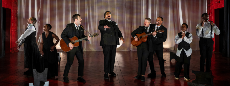 Eight people, two of them with guitars, stand and sing in a line.