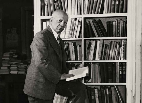 A man holding a book stands in front of a bookshelf.