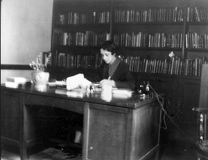 A woman sits at a desk in front of a bookshelf.