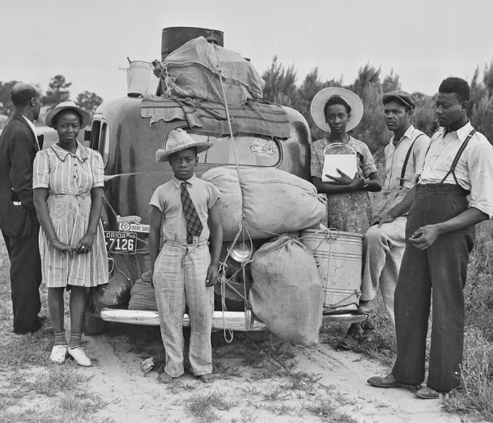 Six family members stand by a fully packed car, with sacks and tins of luggage tied to the back and the roof. On the left there’s an older bald man turned towards the car and away from the camera, a young girl in a straw hat, and an even younger boy in a straw hat and a tie in the center. On the right, two adult men in suspenders and a woman carrying a purse.