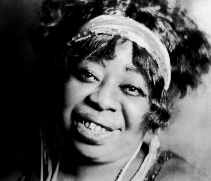 Ma Rainey looks into the camera wearing a cloth headband with tassels hanging behind her ears. She also wears a pearl necklace and a sequined dress. She is smiling warmly with her teeth showing.