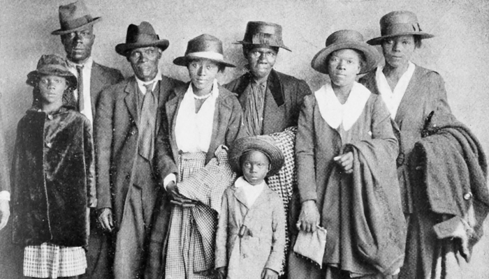 Eight family members wearing traveling hats and coats pose against a wall for a photo. Two suitcases sit in the foreground.