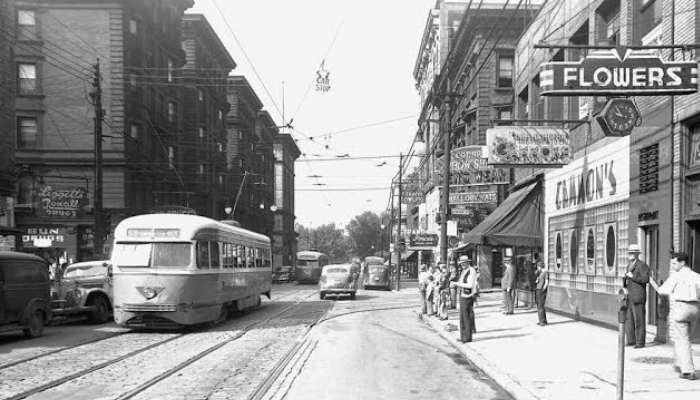 A black-and-white photograph of a street. There is a streetcar in the lefthand corner and a flower shop on the right. Tall brick buildings in the background with other cars in the distance.