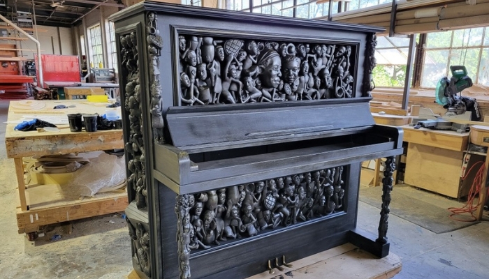 A wooden piano with a dark varnish, several figures are carved into all sides of it. Some faces, some full-body figures.