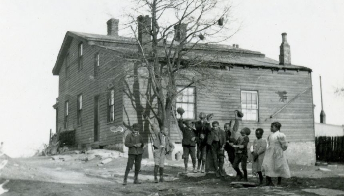 Nine Black children, some waving hats in the air, stand in front of a bare tree outside a wooden house.