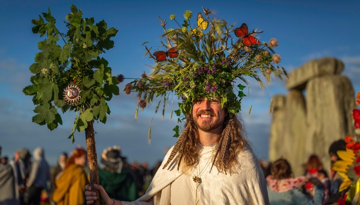 A white man wearing white robes and sporting a tall floral crown made of foliage, flowers, and butterfly figures holds a wooden staff adorned with more flowers. He has feathers in his hair and smiles as the sun lights his face.