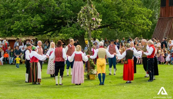 A group of 15 adults wearing brightly colored skirts, slacks, and vests hold hands in a circle around a tall wooden pole adorned with flowers while a crowd of children and adults look on.