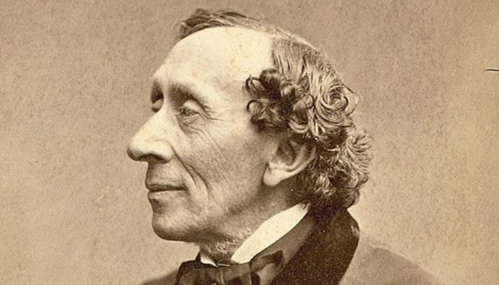 A white man sits in profile posing for a photo portrait. He wears a bow tie and a jacket. His hairline is receding and his curls fall around his ears. He smiles gently as he looks ahead of himself.