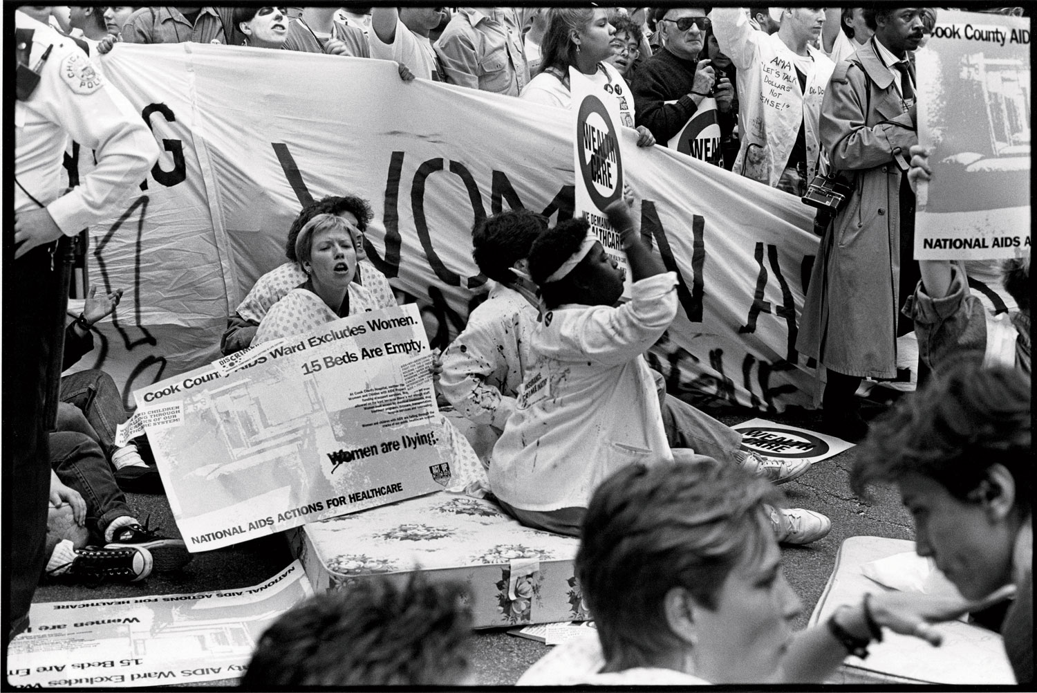 Women sitting on a mattress in the street chant as they hold signs. Behind them, more protesters hold a banner. A police officer is partially visible.
