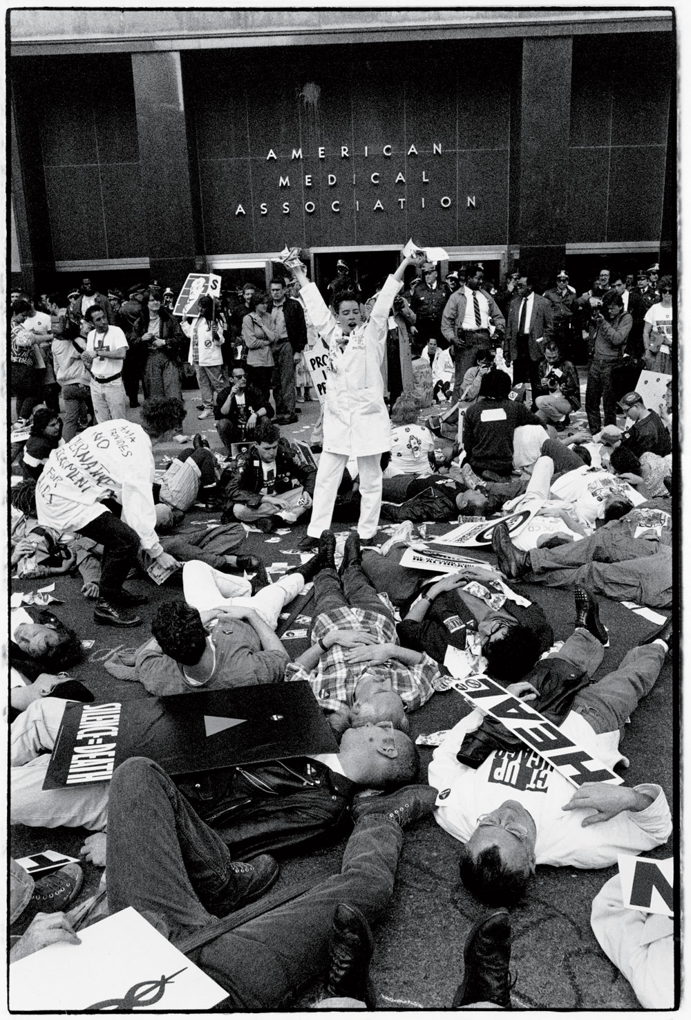 Members of ACT UP stage a die-in. More than a dozen people are visible lying on the ground, with a crowd of onlookers and police officers between them and the AMA building the background. Protesters have signs that read things like “Silence = Death.”