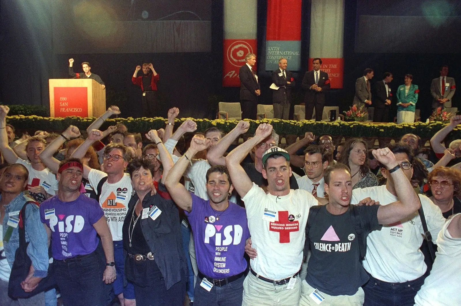 Dozens of protesters stand with their arms around one another, fists in the air as they chant in front of a stage. Their shirts read things like “People with Immune System Disorders” with a pink triangle, and “Silence = Death.” Conference speakers stand awkwardly on the stage behind them.