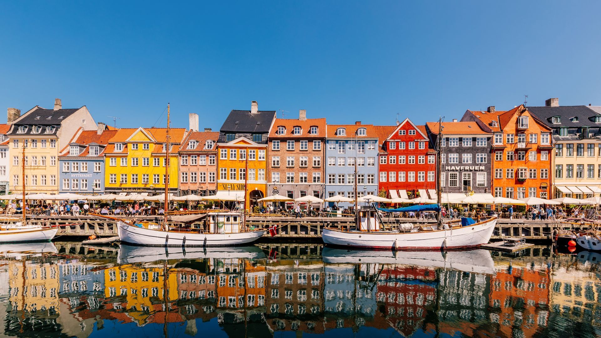 Boats are docked in front of a row of houses in shades of blue, red, orange, yellow, and peach.
