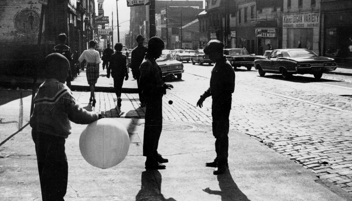Three boys in the foreground play with a ball, three women in the background walk through the street facing away from the camera.