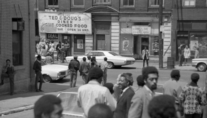 Several people gathered on a street in the foreground, with a restaurant in the background with the sign “Ted & Doug’s Diner/Home Cooked Food.” Several more people are gathered right around the restaurant, and across the street looking towards it.