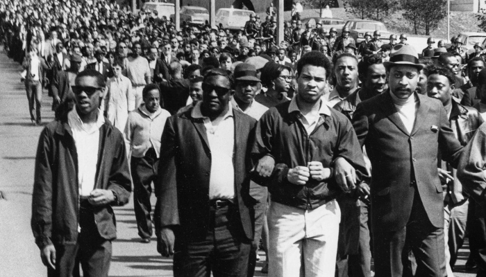 Several hundred people fill the street and march toward the camera, walking arm and arm. At the front of the photo there are four men - two with sunglasses, one with an afro, and one with a hat, all in collared shirts and suits.