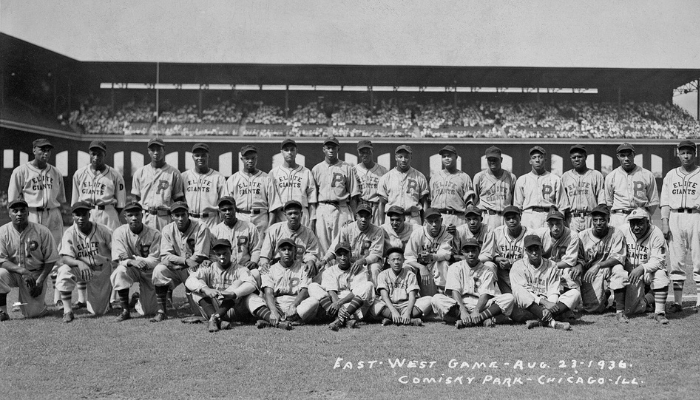 35 Black players arranged in three rows on the field with the stadium full of fans behind them. Their uniforms say P for Pittsburgh Crawfords or Elite Giants for the Chicago American Giants.