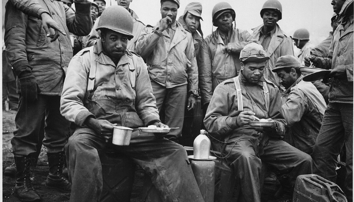 A few Black soldiers sit and eat on the sand. Many others stand behind them noticing the camera.
