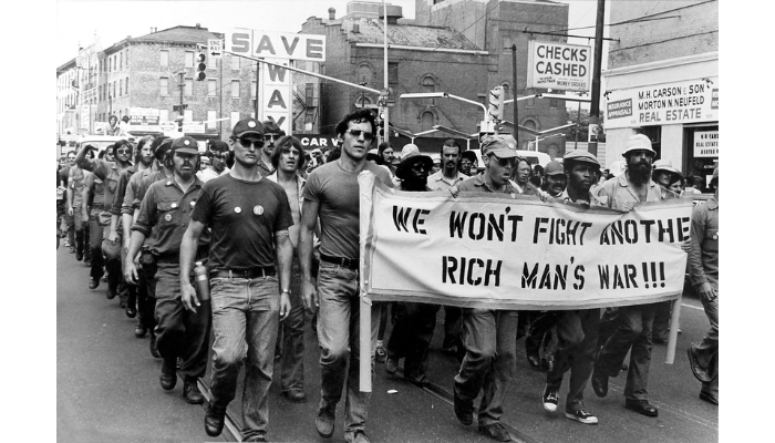 A densely packed group of both Black and white men march in a street protest. They hold up a sign that reads “We won’t fight another rich man’s war.” Storefronts for real estate offices and a checks cashed station are in the background.