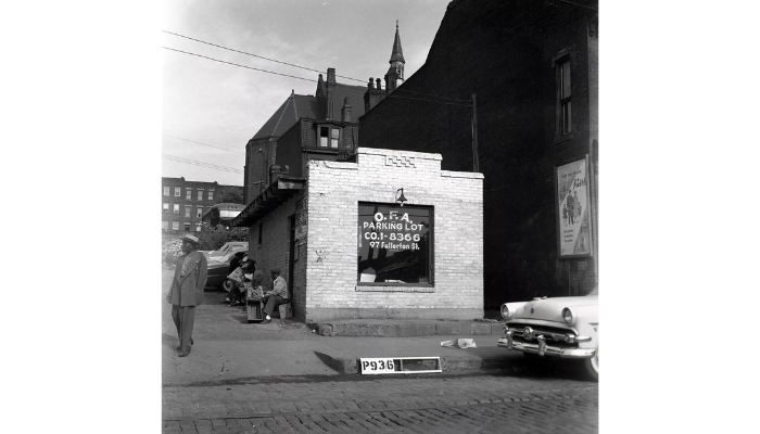 A brick building with a window that reads: ‘O.F.A. PARKING LOT - CO. 1-8366, 97 Fullerton Street.’ Several men sit in the shade on the side of the building.