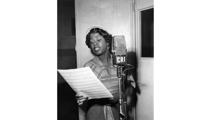 A Black woman sings in front of a microphone, carrying sheet music in her left hand. She wears a striped dress and a hat.
