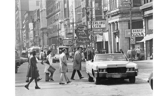 Several individuals cross a busy street, a light-colored car is in the foreground driving towards the camera. In the background are signs for a furniture store called Sterling Lite, A flower shop, and a loan store.