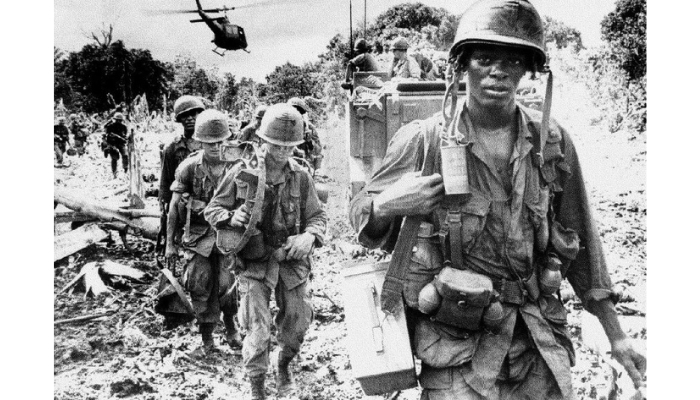 Several men march toward the camera carrying equipment on their backs. There is a Black soldier in the foreground looking at the camera. There are trees in the background and a helicopter flying overhead.