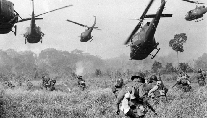 Five helicopters fly and fire as soldiers navigate tall grass towards the treeline.