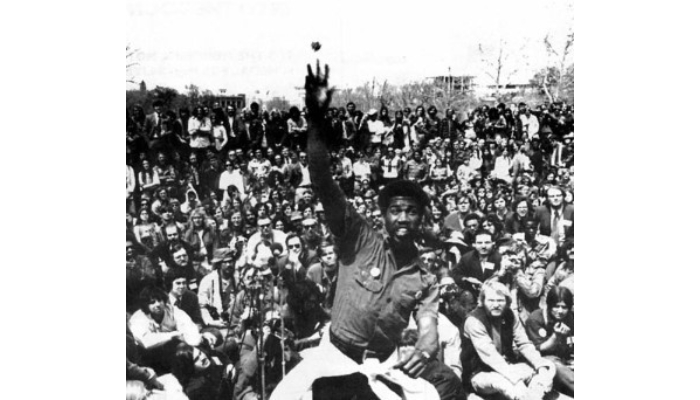 A Black veteran hurls his medal in the direction of the camera while a large crowd of people sit and stand behind him, looking on.