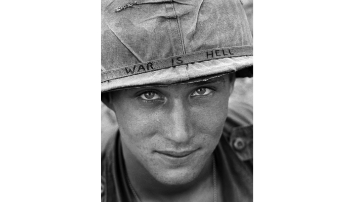 A close-up shot of a soldier with a slight smile, looking directly at the camera. He wears a helmet, the band of which says War is Hell.
